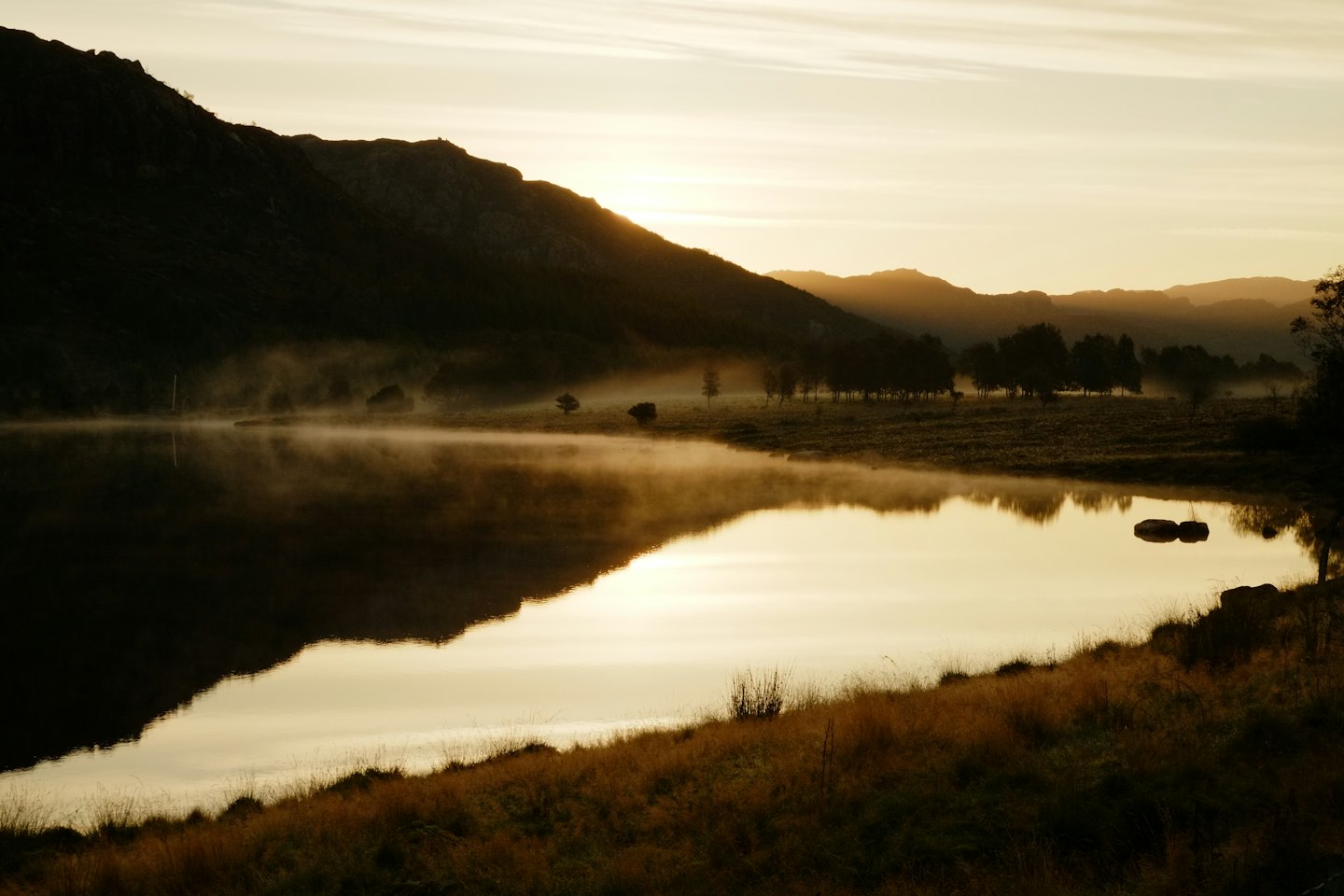 Misty lake reflecting golden sunrise light behind mountains.